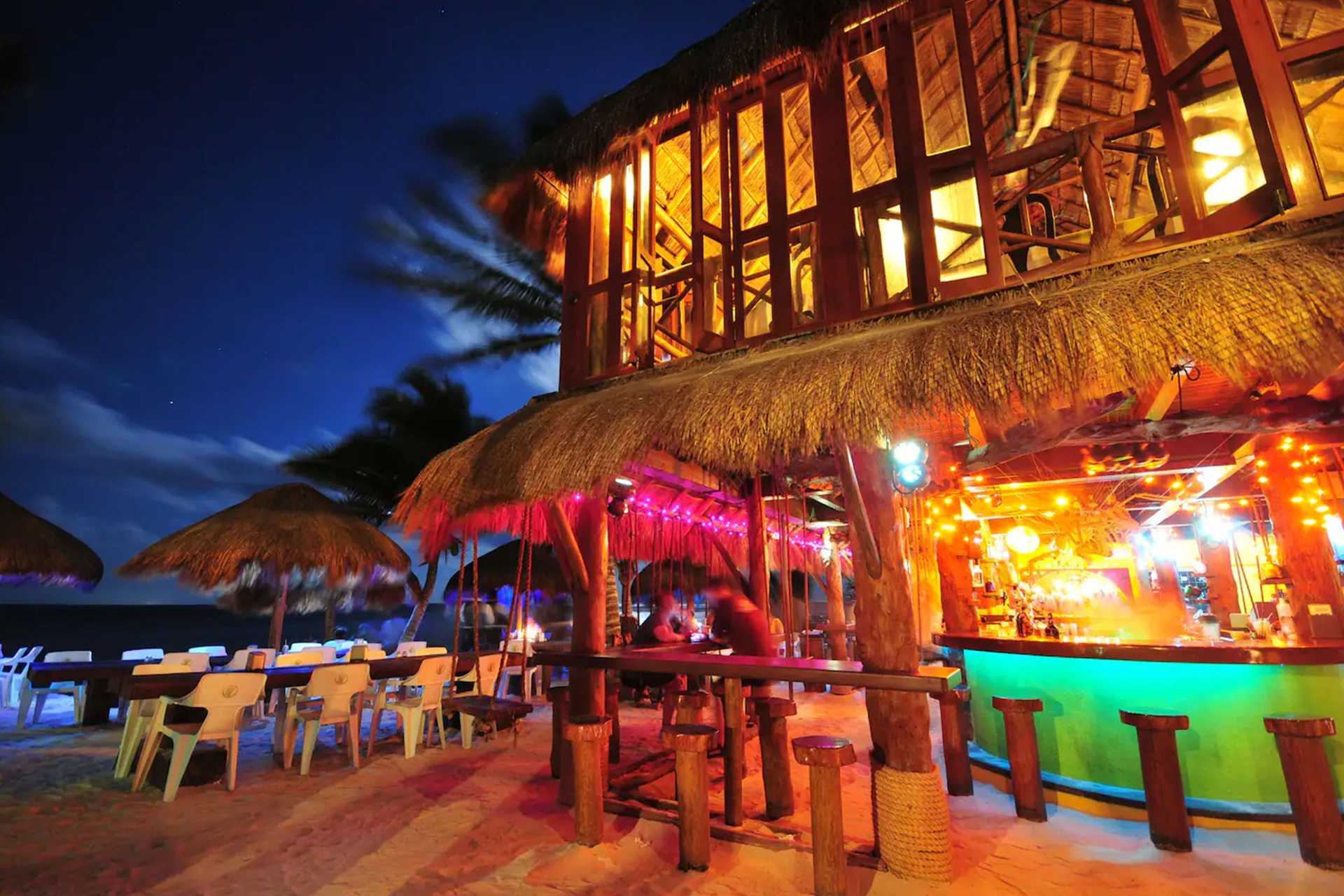 La Buena Vida beachfront restaurant at night with thatched palapa roof, colorful bar lights, and sandy dining area under palm trees